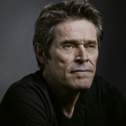 Willem Dafoe looks calmly into the camera from a side angle - the right side of his face more visible