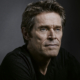 Willem Dafoe looks calmly into the camera from a side angle - the right side of his face more visible